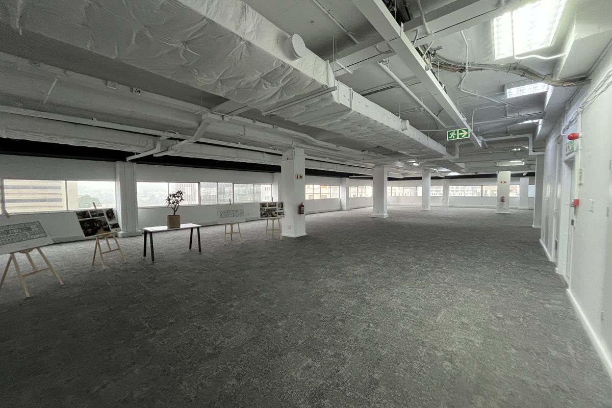 A spacious, open-plan office area featuring exposed industrial ceilings, large windows providing natural light, and neutral carpeted flooring throughout.