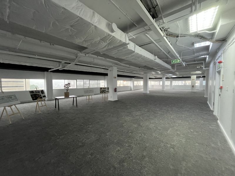 A spacious, open-plan office area featuring exposed industrial ceilings, large windows providing natural light, and neutral carpeted flooring throughout.