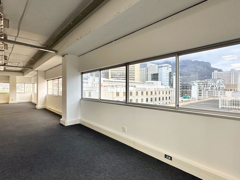 A spacious, open-plan office area featuring large windows with city and mountain views, carpeted flooring, and exposed ceiling infrastructure.