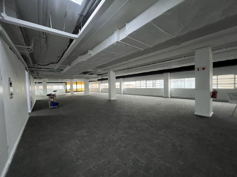 A spacious, open-plan office area featuring exposed ceiling ducting, carpeted flooring, and large windows providing ample natural light.