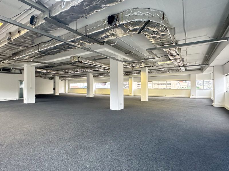 Spacious open-plan office area featuring exposed industrial-style HVAC ducting, recessed lighting, carpeted flooring, and large windows providing ample natural light.