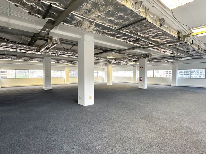 A spacious, open-plan office area featuring exposed ceiling ducting, carpeted flooring, and large windows providing ample natural light.