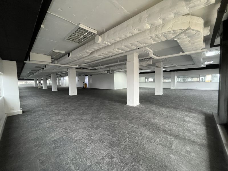 Spacious open-plan office area featuring exposed ceiling ductwork, structural columns, carpeted flooring, and large windows providing ample natural light.