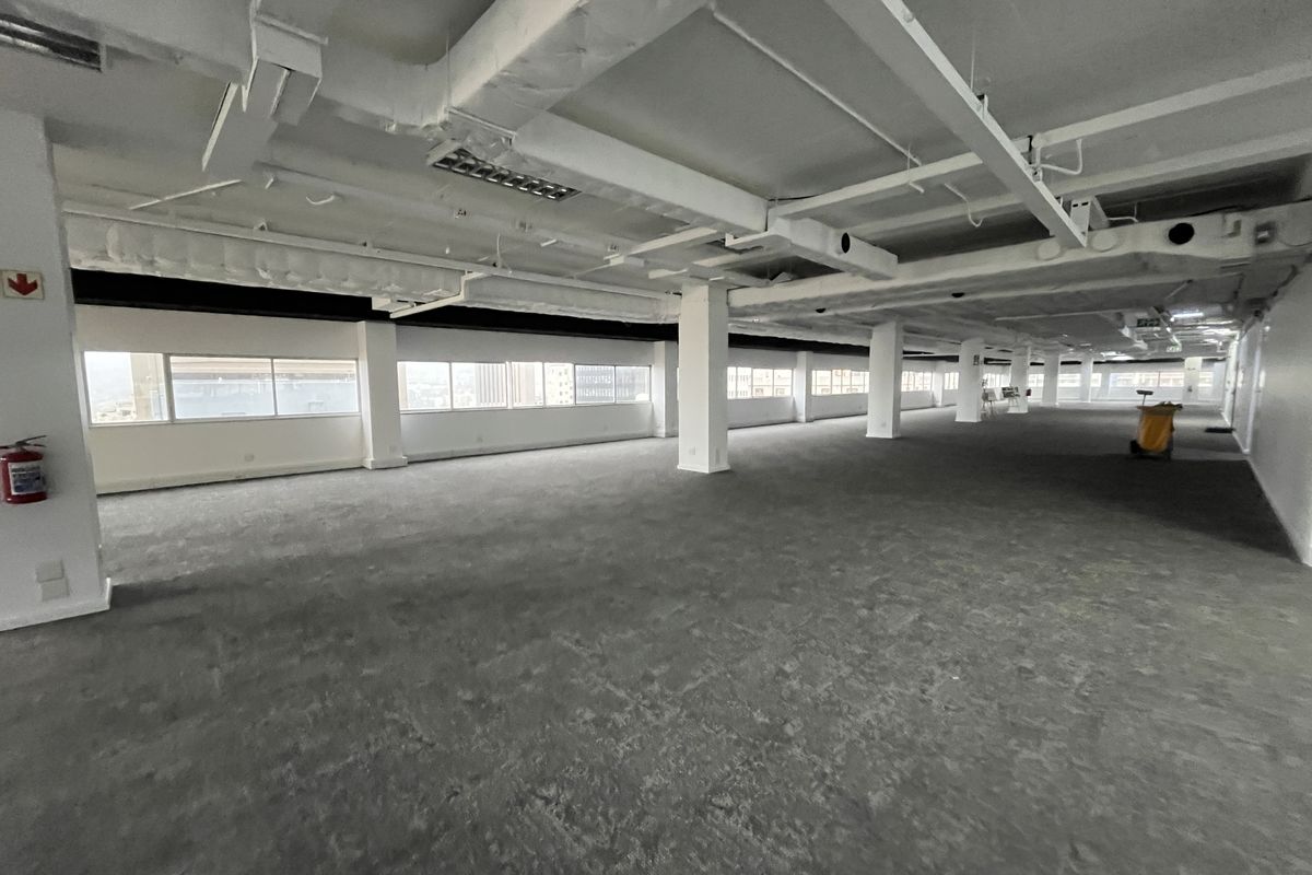 A spacious, open-plan office area featuring exposed industrial ceilings, large windows providing natural light, and neutral carpeting throughout.