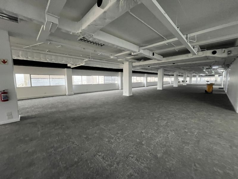 A spacious, open-plan office area featuring exposed industrial ceilings, large windows providing natural light, and neutral carpeting throughout.