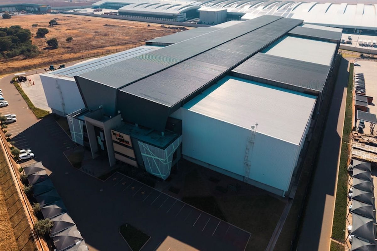 Aerial view of a large industrial warehouse facility featuring a modern metal roof, extensive parking area, and secure perimeter fencing.