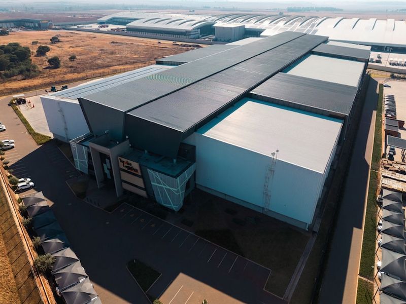 Aerial view of a large industrial warehouse facility featuring a modern metal roof, extensive parking area, and secure perimeter fencing.