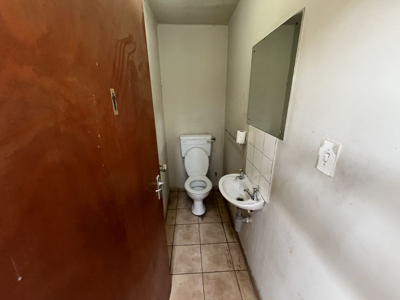 A functional industrial-style restroom featuring a toilet, wall-mounted basin, tiled flooring, and a wall mirror, requiring cosmetic updates to the wall finishes.