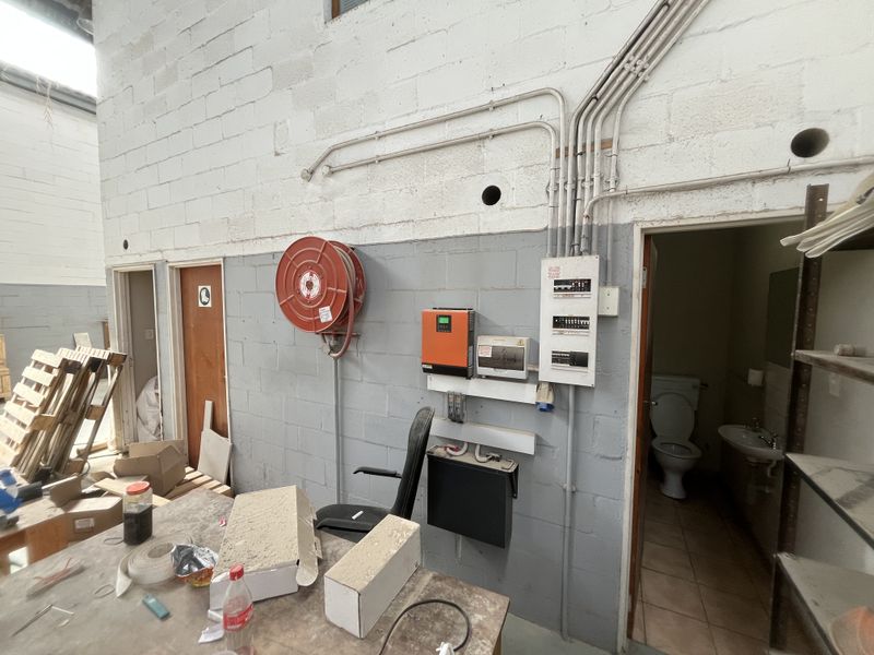 Industrial utility space featuring exposed conduit, fire safety equipment, electrical infrastructure, and an adjacent small restroom facility with basic sanitary fixtures.