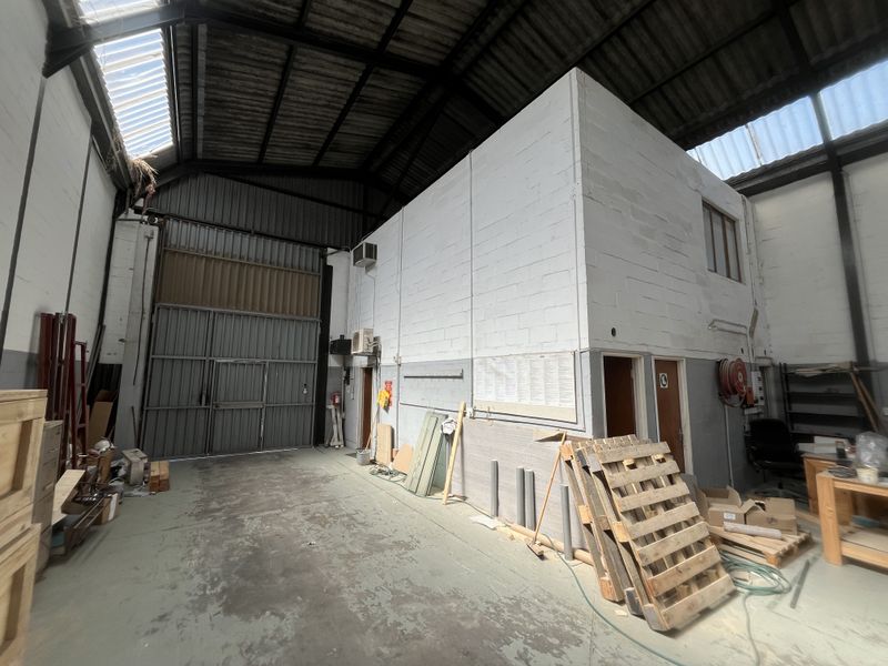 Spacious industrial warehouse unit featuring high ceilings with exposed steel trusses, skylights, a large roller shutter door, and a mezzanine level.