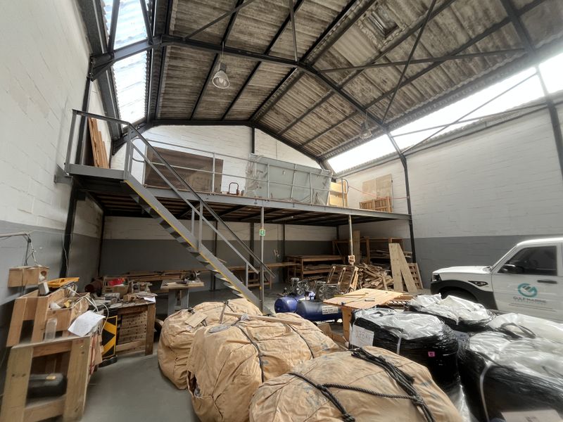 Industrial warehouse space featuring a mezzanine level, exposed steel roof trusses, translucent roof panels, and concrete flooring, suitable for storage or workshop operations.