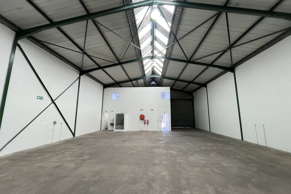 Spacious industrial warehouse unit featuring high ceilings with exposed steel trusses, natural skylight illumination, concrete flooring, and a large roller shutter door.