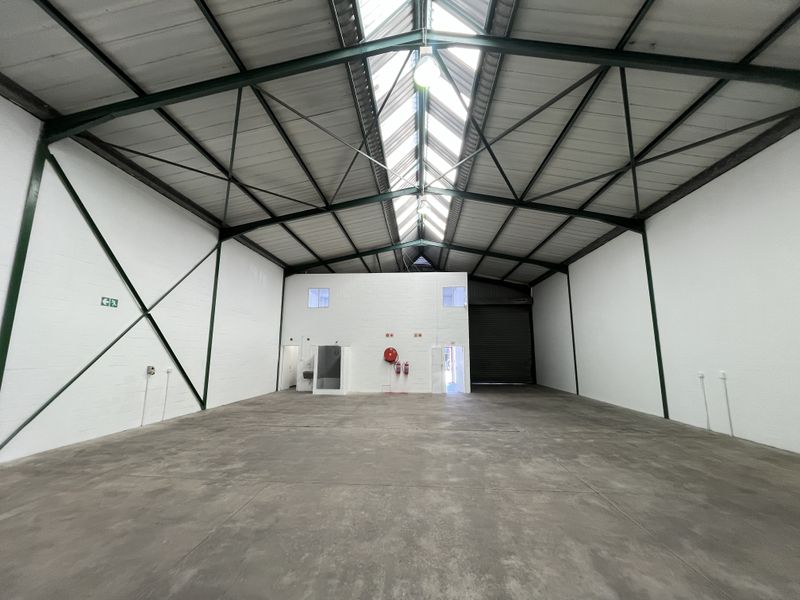 Spacious industrial warehouse unit featuring high ceilings with exposed steel trusses, natural skylight illumination, concrete flooring, and a large roller shutter door.
