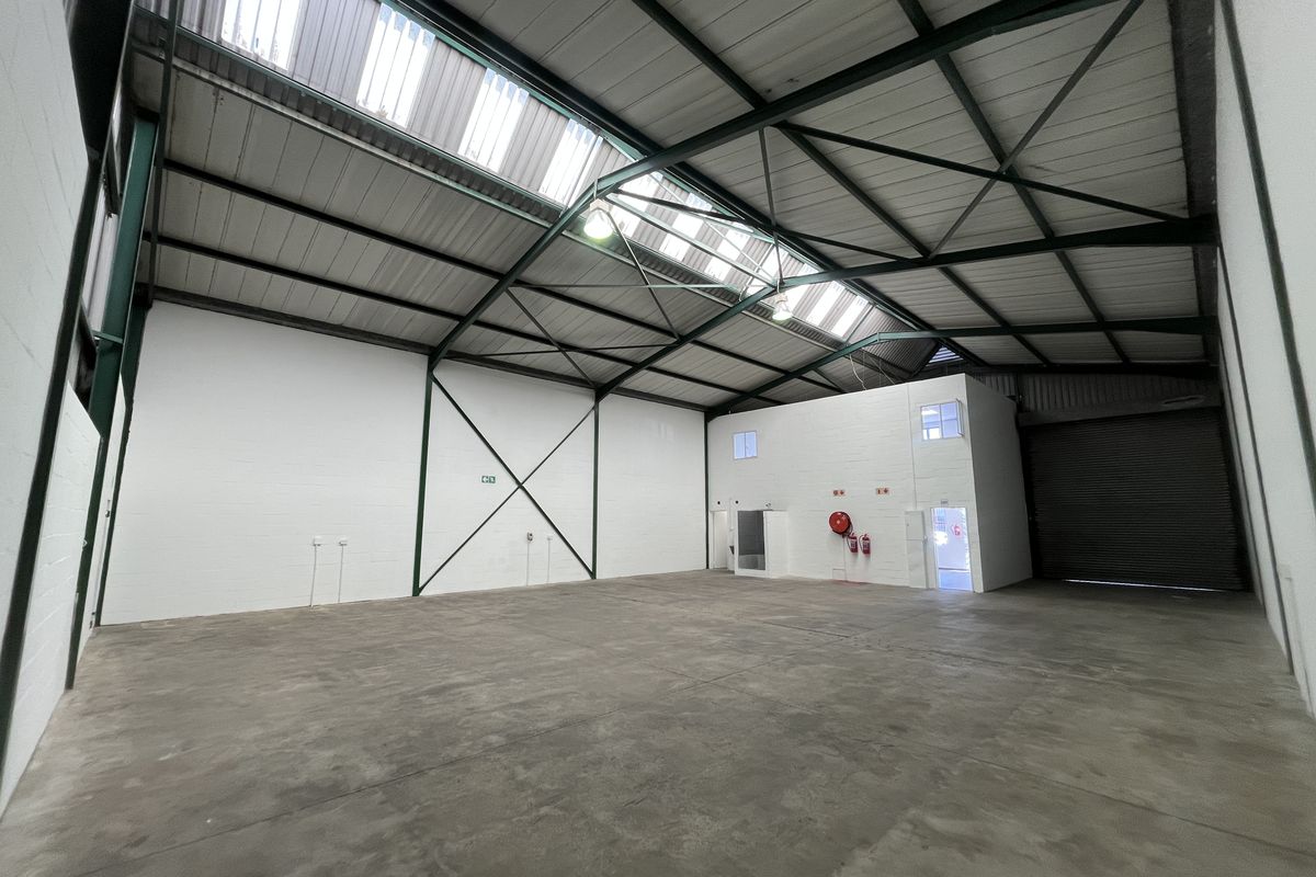 Spacious industrial warehouse unit featuring high ceilings with translucent roof panels, concrete flooring, a large roller shutter door, and an integrated mezzanine office area.