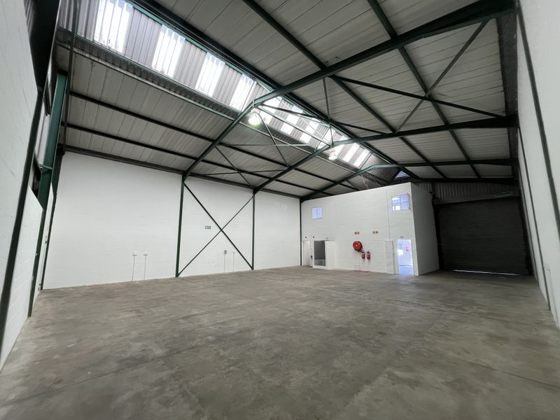 Spacious industrial warehouse unit featuring high ceilings with translucent roof panels, concrete flooring, a large roller shutter door, and an integrated mezzanine office area.