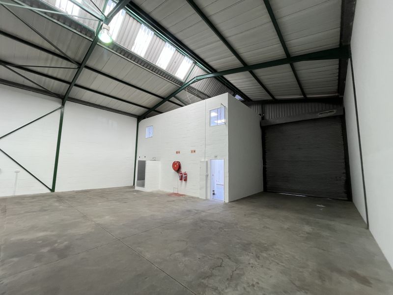 Spacious industrial warehouse unit featuring high ceilings with exposed steel trusses, skylights, concrete flooring, a roller shutter door, and an internal office structure.