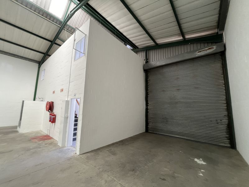 Industrial warehouse space featuring high ceilings with exposed steel trusses, a large roller shutter door, and a self-contained internal office or utility block.