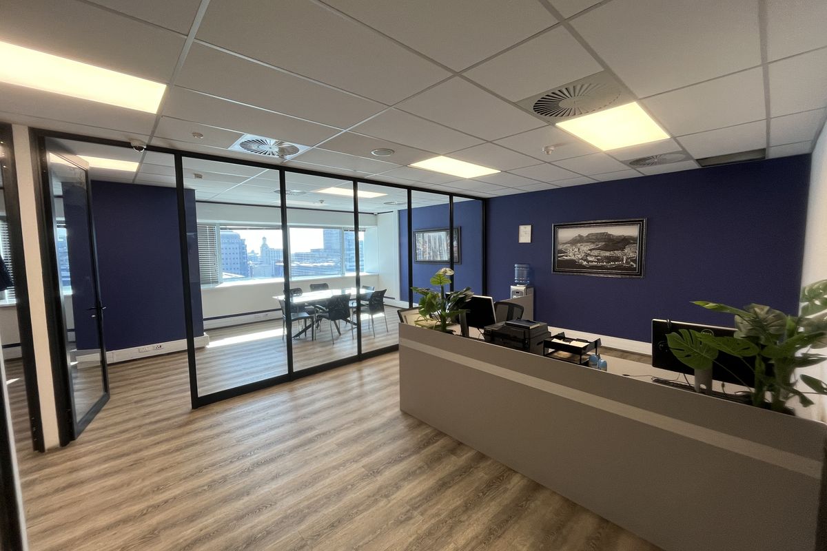 Modern office space featuring a glass-partitioned meeting room, wood-look laminate flooring, and a professional reception area with recessed ceiling lighting.