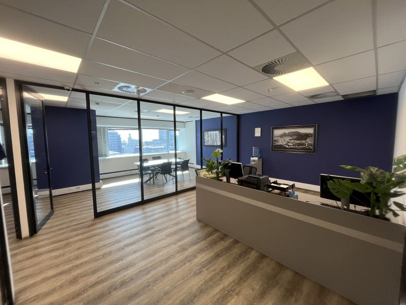 Modern office space featuring a glass-partitioned meeting room, wood-look laminate flooring, and a professional reception area with recessed ceiling lighting.