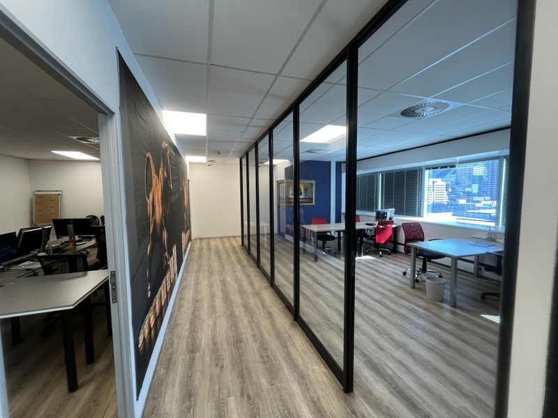 Modern office space featuring wood-look laminate flooring, suspended ceiling with recessed lighting, and a sleek black-framed glass partition wall.