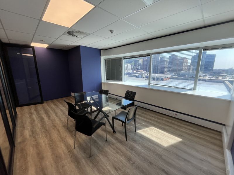 A modern office space featuring laminate flooring, a suspended ceiling with recessed lighting, and large windows offering an urban city view.