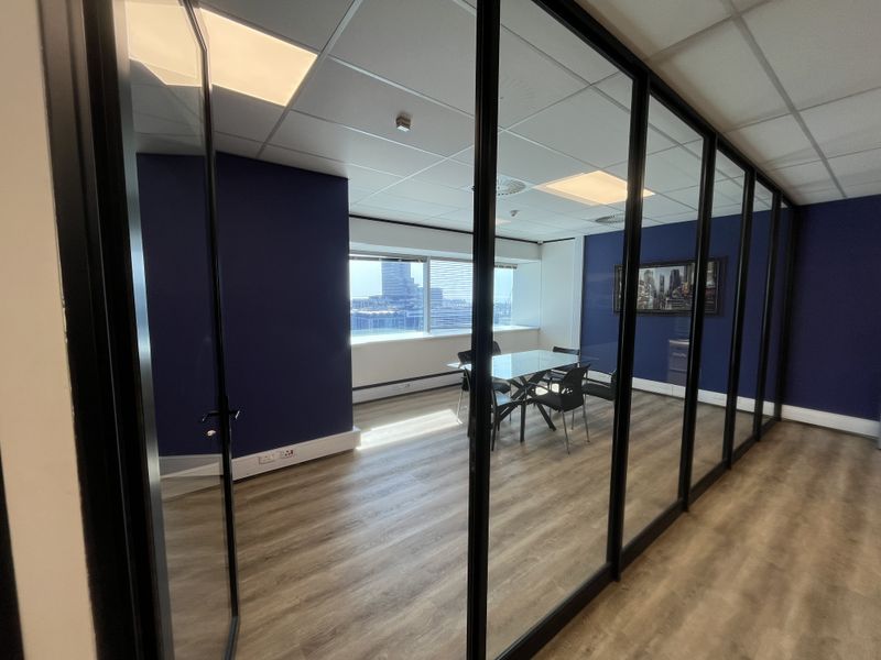 A modern office space featuring glass partition walls, wood-look laminate flooring, and recessed lighting, offering a professional environment with natural light.