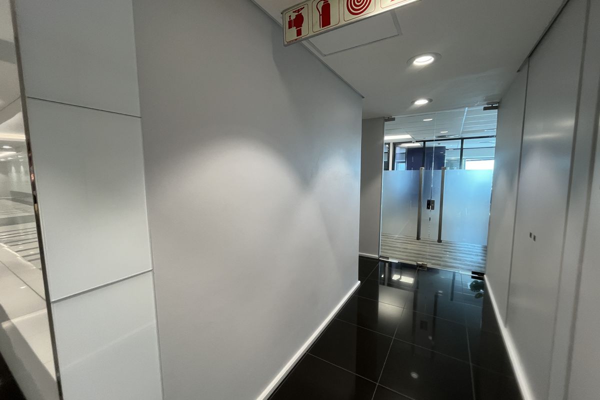 A clean, modern office hallway featuring polished dark tiles, recessed lighting, and a glass entrance door with clear fire safety signage.