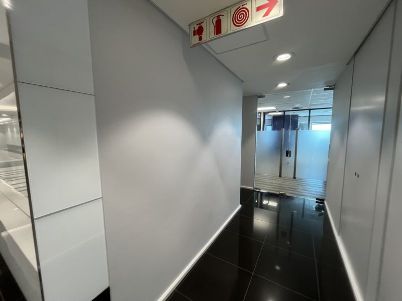 A clean, modern office hallway featuring polished dark tiles, recessed lighting, and a glass entrance door with clear fire safety signage.