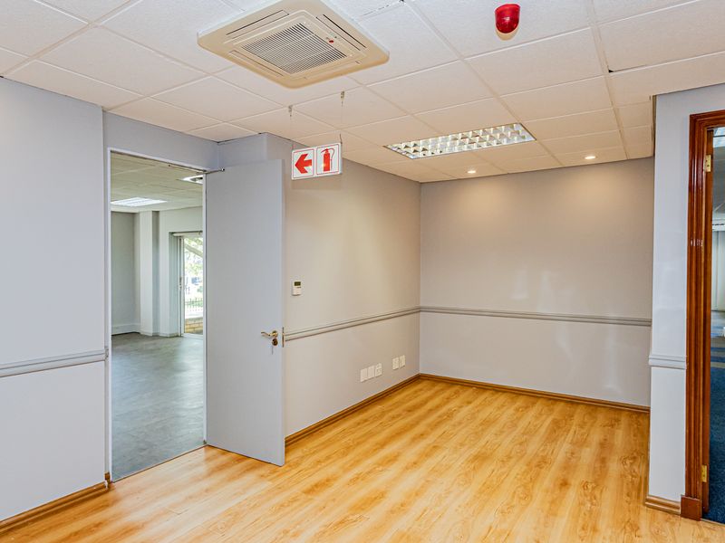 A bright, empty office space featuring laminate flooring, a suspended ceiling with recessed lighting, integrated air conditioning, and a clear view into adjacent office areas.