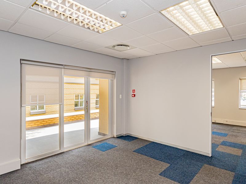 A bright, empty office space featuring carpet tiles, a suspended ceiling with recessed lighting, and a sliding glass door leading to an outdoor area.