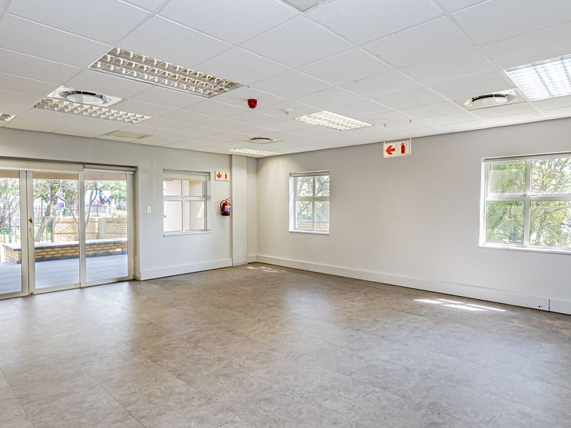 A spacious, well-lit open-plan office area featuring a suspended ceiling, vinyl flooring, and a small built-in kitchenette with direct access to an outdoor patio.