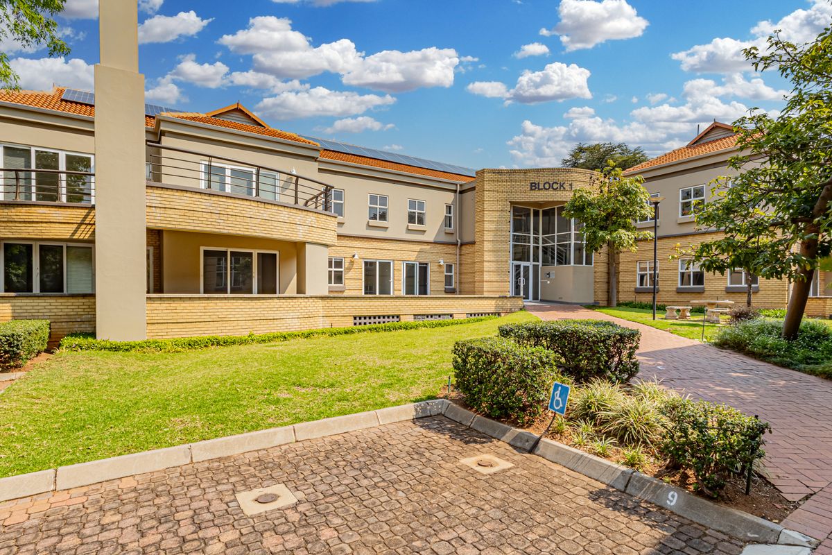 A well-maintained office building exterior featuring a brick facade, tiled roof, balcony, and landscaped garden area with paved access.