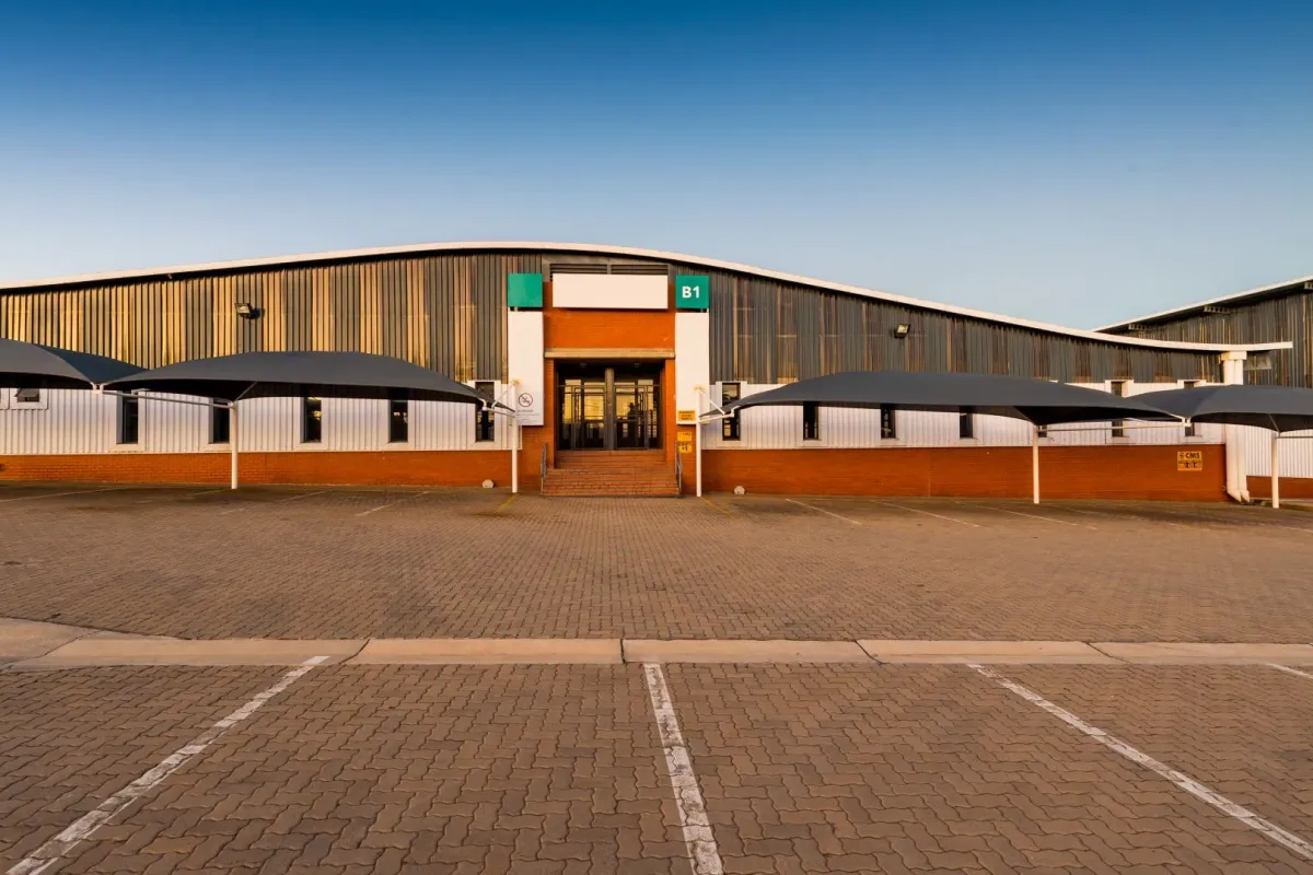 Front elevation of an industrial warehouse facility featuring corrugated metal cladding, a paved parking area with shade sails, and a central glass entrance.
