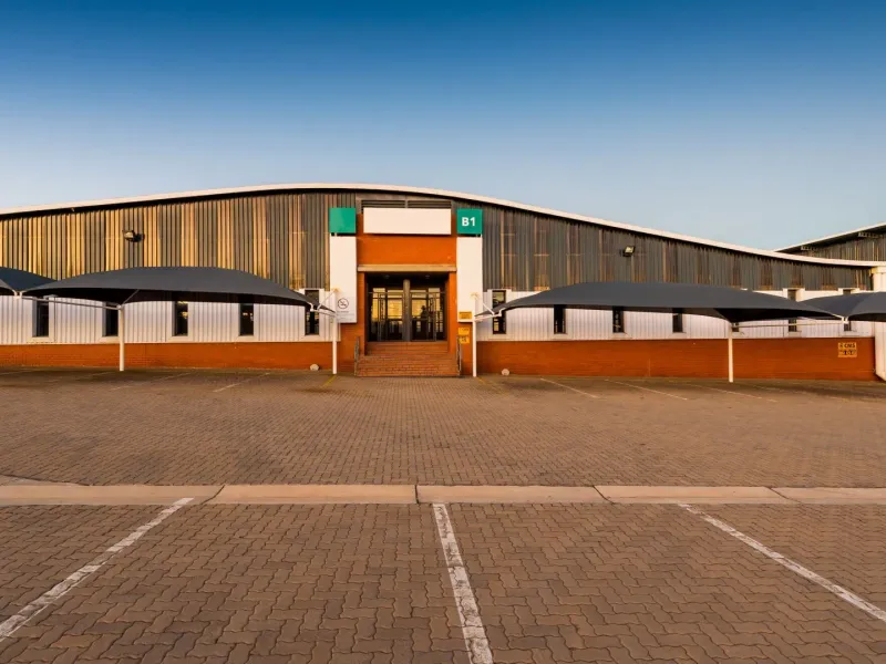 Front elevation of an industrial warehouse facility featuring corrugated metal cladding, a paved parking area with shade sails, and a central glass entrance.