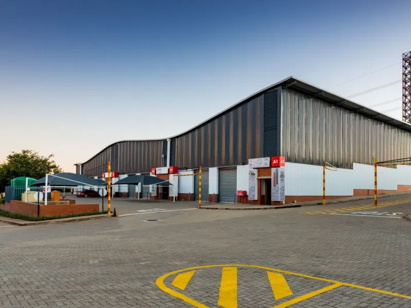 Modern industrial warehouse facility featuring corrugated metal cladding, multiple roller shutter doors, paved yard area, and dedicated shade cloth parking bays.
