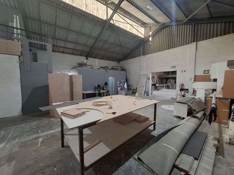 Spacious industrial workshop area featuring high ceilings with exposed steel trusses, concrete flooring, and painted brick walls, suitable for manufacturing or storage operations.