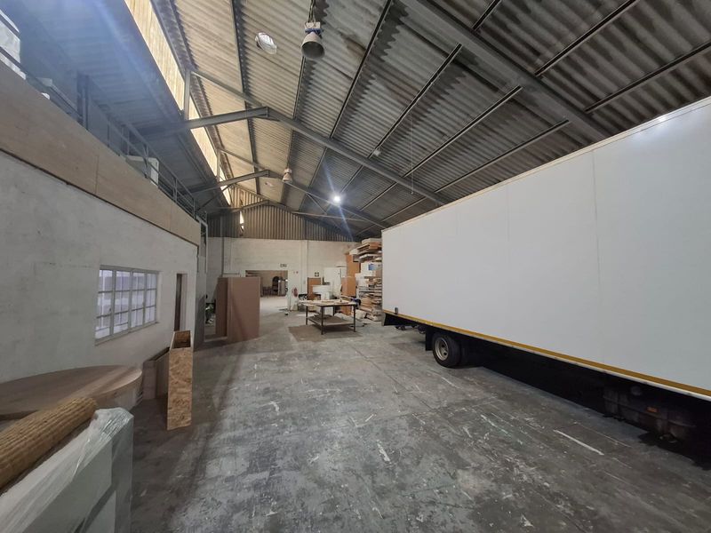 Spacious industrial warehouse area featuring high ceilings with exposed steel trusses, concrete flooring, and ample natural light through multi-pane windows.