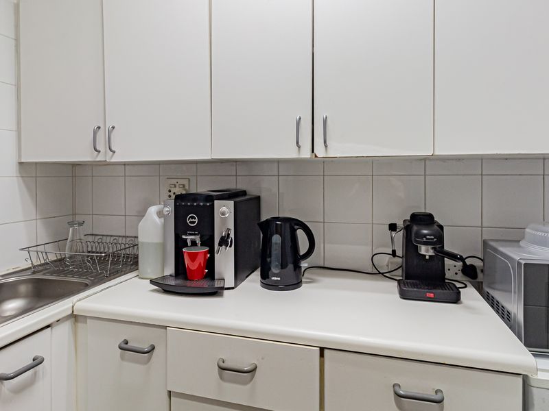 A functional office kitchenette featuring white built-in cabinetry, a laminate countertop, and a stainless steel sink with a tiled backsplash.