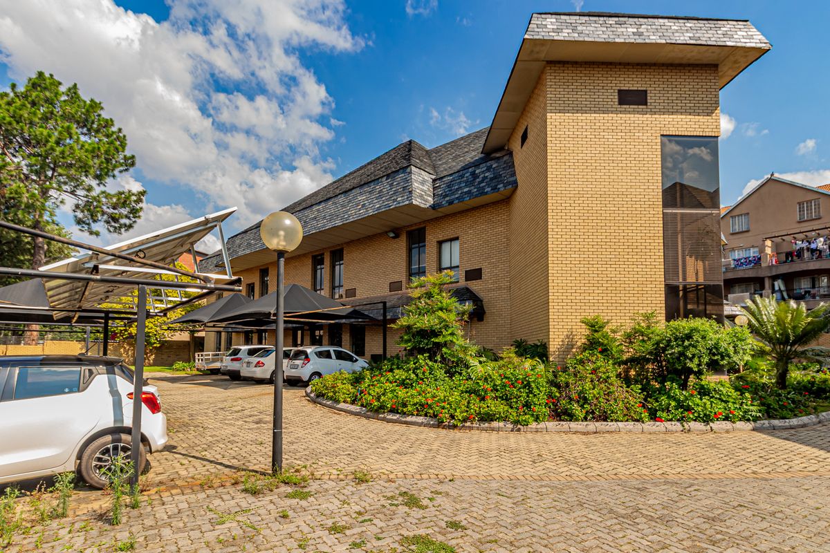 Front exterior view of a multi-story brick office building featuring a paved parking area with covered carports and established garden landscaping.