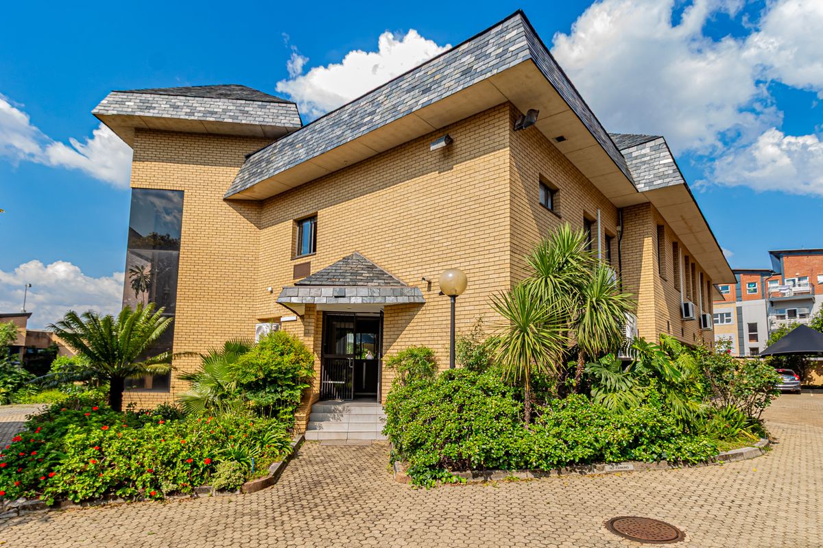 A professional exterior view of a multi-story brick office building featuring a slate roof, paved driveway, and established garden landscaping.