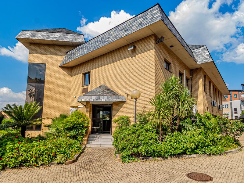 A professional exterior view of a multi-story brick office building featuring a slate roof, paved driveway, and established garden landscaping.