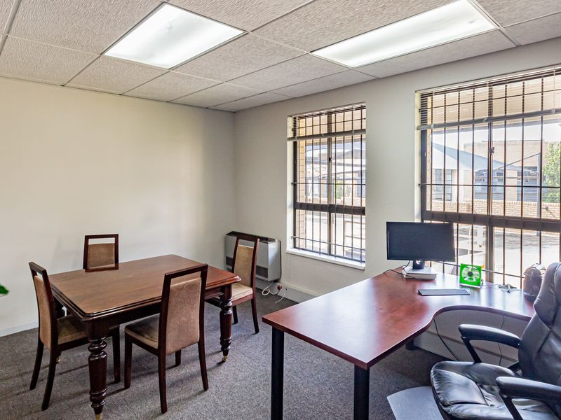 A functional office space featuring carpeted flooring, suspended ceiling with recessed lighting, and windows fitted with security bars.