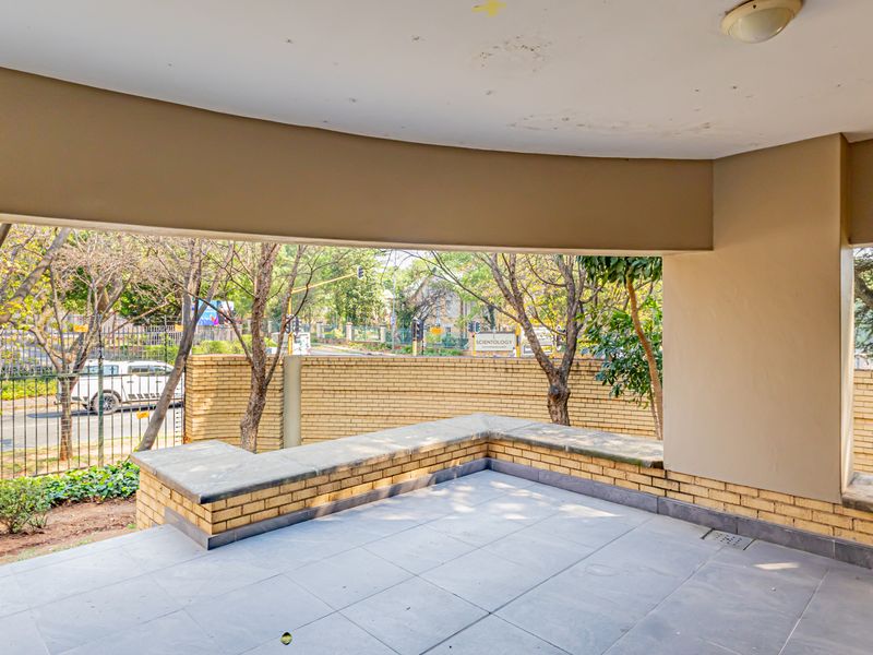 A covered outdoor patio area featuring built-in brick seating, tiled flooring, and views of the surrounding street and boundary wall.
