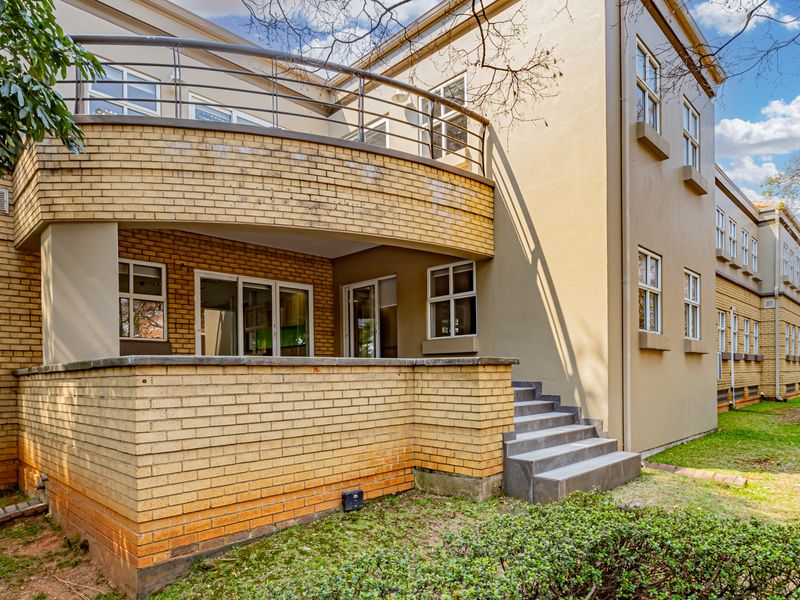 Rear exterior view of the office building featuring a brick facade, a curved balcony with metal railings, and concrete steps leading to the garden area.
