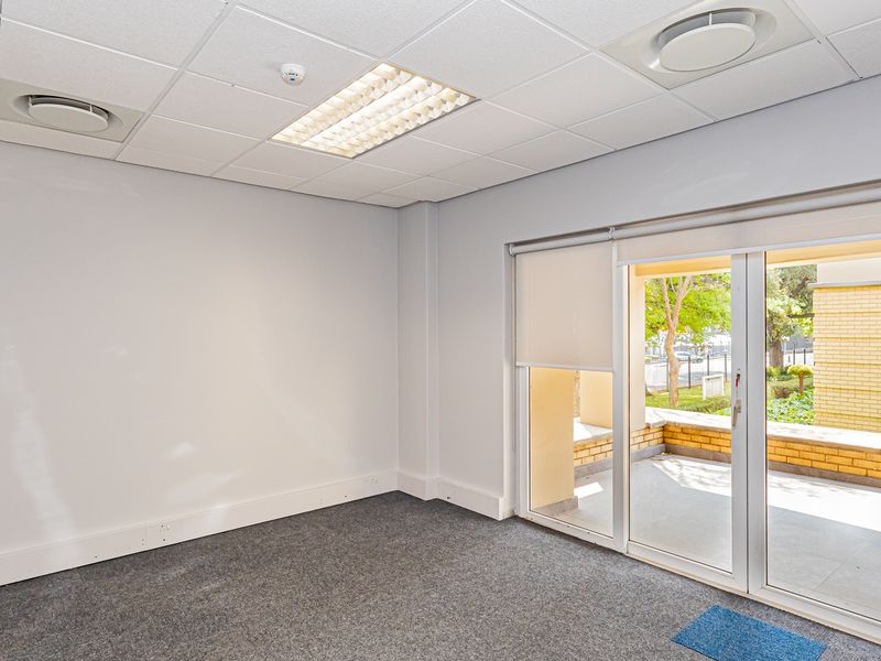 A bright, empty office space featuring a suspended ceiling, recessed lighting, carpeted flooring, and a sliding glass door leading to an outdoor patio area.