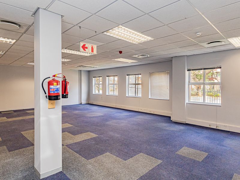 A spacious, vacant open-plan office area featuring carpeted flooring, suspended ceilings with recessed lighting, and large windows providing ample natural light.