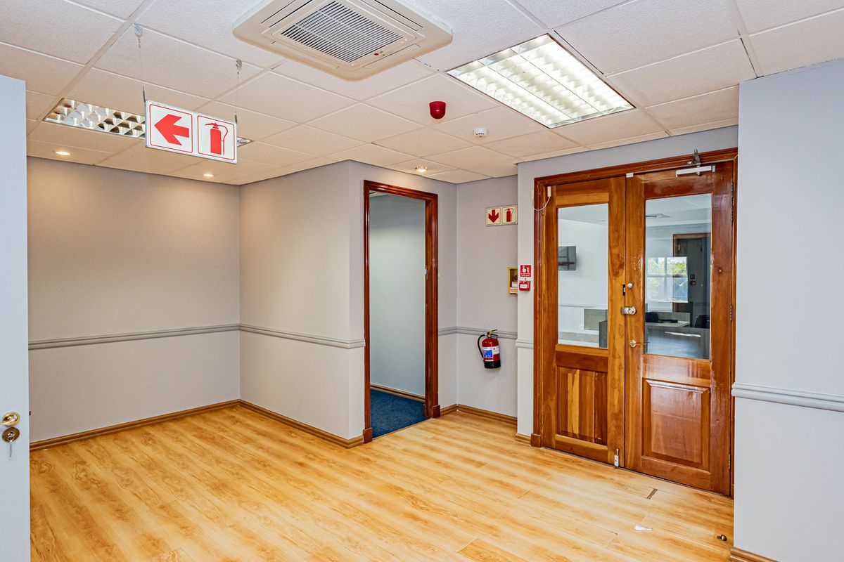 A clean, empty office space featuring laminate flooring, a suspended ceiling with recessed lighting, and wooden double doors leading to an adjacent room.