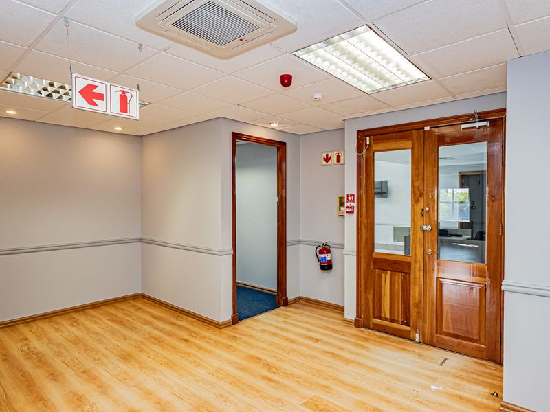 A clean, empty office space featuring laminate flooring, a suspended ceiling with recessed lighting, and wooden double doors leading to an adjacent room.