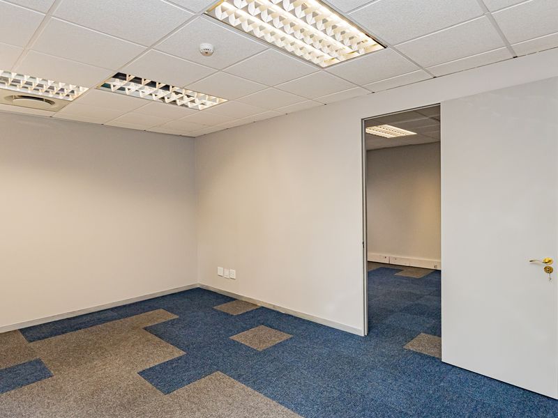 A clean, empty office space featuring neutral walls, carpet tile flooring, and recessed lighting, with an adjacent room visible through the doorway.