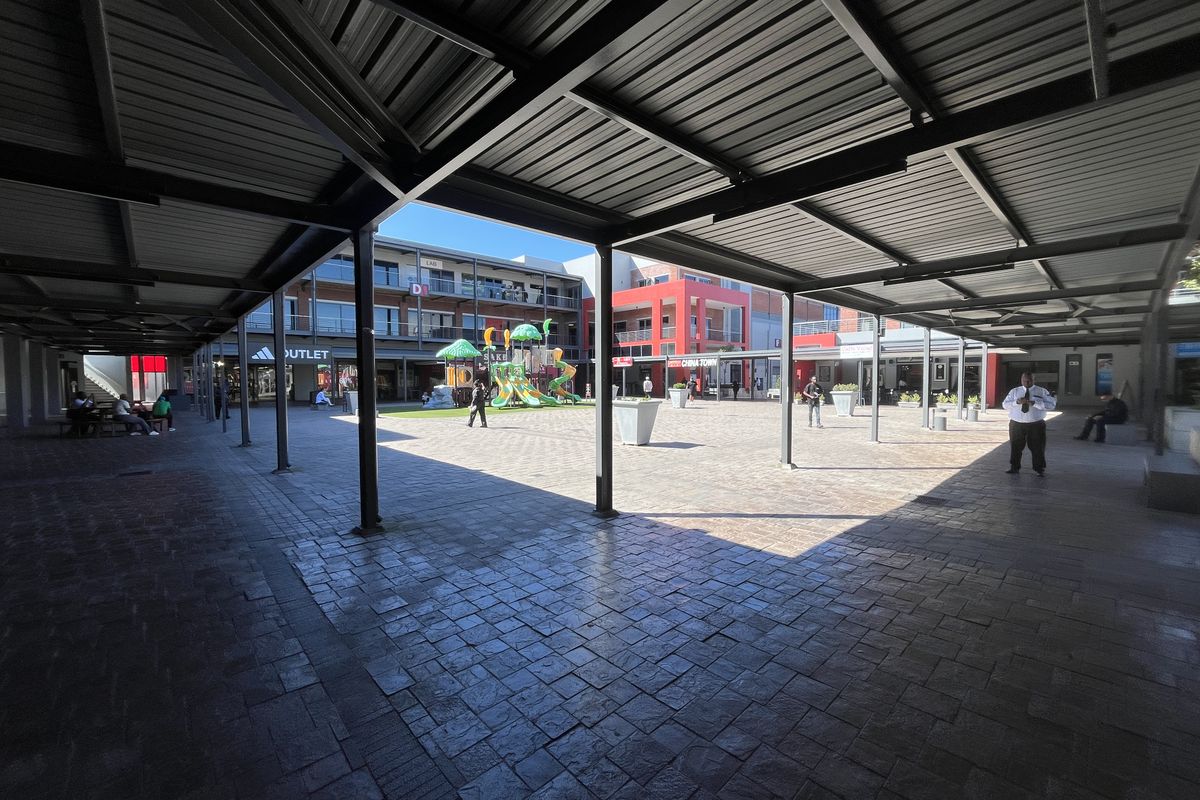 A spacious covered outdoor retail plaza featuring paved flooring and a modern steel roof structure, providing a sheltered walkway between commercial storefronts.